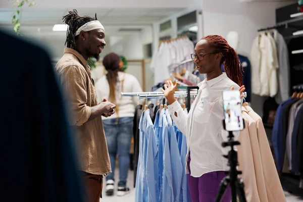 Clothing store assistant giving interview to fashion blogger while recording promotional video on smartphone. African american influencer and shop worker advertising outfit for social media