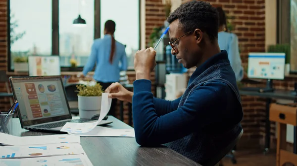 アフリカ系アメリカ人の男性のブレインストーミングのアイデアは 完全な意思決定を行うために統計と紙やラップトップを使用して 新しいビジネスビジョンを作成します オフィスにおける新しい研究計画について考える会社のアナリスト — ストック写真