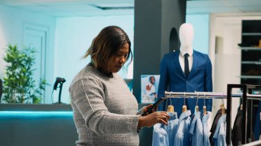 Afro-Amerikalı bir kadın web sitesi hisselerine bakıyor, giyim mağazasında internet uygulamasından kıyafet bulmaya çalışıyor. Genç müşteri akıllı telefonu alışveriş merkezindeki favori ürünleri almak için kullanıyor..