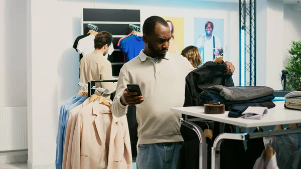 Male shopper looking at online website to find clothes, buying new fashionable merchandise in clothing store. Person with smartphone checking stock of formal or casual wear, shopping mall.