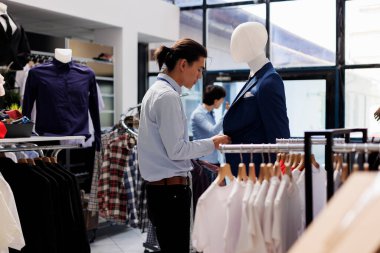 Asyalı işçiler gündelik kıyafetlere bakıyor, mağaza açılmadan önce şık kıyafetler hazırlıyorlar. Resmi tişört giyen bir işçi, modern butikte moda ürünlerle çalışıyor. Moda konsepti