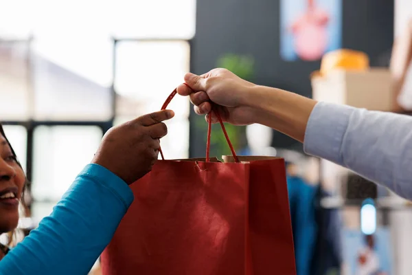 在现代精品店 员工在为新衣橱购买时尚服装后 将红色纸袋送给顾客 非洲裔美国妇女在服装店购买休闲装 — 图库照片