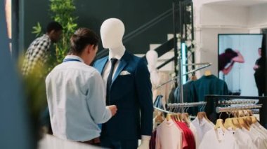 Şık bir adam resmi takım elbiseyle mankeni kontrol ediyor, mağaza açılmadan önce şık kıyafetler ayarlıyor. Modern butikte Asyalı çalışanlar moda ürünler ve aksesuarlarla çalışıyor. Moda konsepti