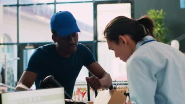 Asyalı işçi paket teslimatı raporunu imzalıyor, modern butikte Afro-Amerikan kuryesiyle kıyafet dağıtımını tartışıyor. Mağaza müdürü alışveriş merkezinde müşteri siparişleriyle dolu kutular hazırlıyor