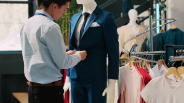 Asyalı işçi mavi takım elbiseli manken ayarlıyor, mağaza açılmadan önce şık kıyafetleri kontrol ediyor. Alışveriş merkezinde son moda ürünler ve aksesuarlarla çalışan şık bir çalışan. Moda konsepti