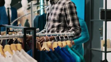 Gündelik giysiler için alışveriş yapmak, modern butikte kumaş ürünlerini analiz etmek. Afro-Amerikalı bir adam yeni moda koleksiyonu olan askılara bakıyor, temel kıyafetleri kontrol ediyor.