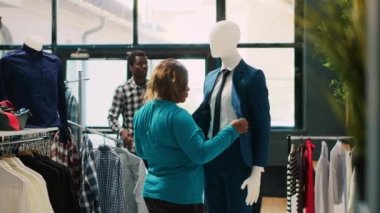 Afro-Amerikalı kadın mavi takımı kontrol ediyor, elbise mağazasında kumaş analizi yapıyor. Şık bir alıcı, modaya uygun mallar için alışveriş yapıyor, modern butikten şık gömlekler alıyor. Moda konsepti