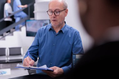 Hastane lobisinde sıhhiyeye danışmaya başlamadan önce sağlık sigortası belgelerini imzalayan kıdemli bir adam. Resepsiyonist hasta kayıtlarını yönetiyor ve takip randevularını ayarlıyor.