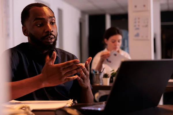 プロジェクトマネージャーは テレビ会議で同僚とスタートアップについて話し合い 夜はオフィスでインターネットの電話に答える企業の労働者 アフリカ系アメリカ人男性オンライン会議コンピュータアプリを使用して — ストック写真