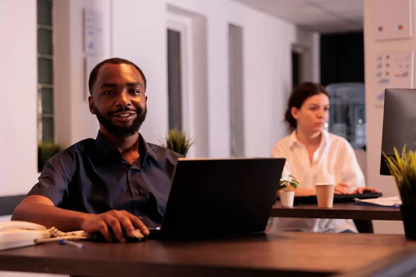 同僚のスペースでノートパソコンで働く従業員 カメラを見て 夜に職場のデスクに座っている企業の労働者 コンピュータを使ってアフリカ系アメリカ人の笑顔を — ストック写真