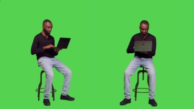 Erkek model yeşil ekranda dizüstü bilgisayarla çalışıyor. Stüdyoda sandalyede oturuyor ve bilgisayardaki internet sitesine bakıyor. Stüdyodaki kablosuz bilgisayarda internet sayfasını kontrol eden genç bir yetişkin.