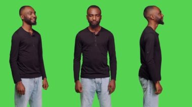Genç erkek manken kamerada şık poz veriyor, yeşil ekran arka planında duruyor. Kot pantolon ve gömlek giyen, kendine güveni olan, stüdyoda pozitif yüz ifadeleri takınan sıradan bir insan..