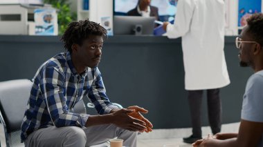 African american men sitting in waiting area at facility and talking about medical insurance support. Male patients waiting to attend examination, discussing about treatment or medicine.