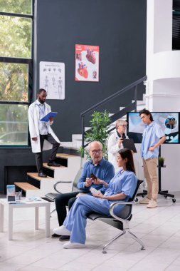 Hastane bekleme odasında sağlık kontrolü sırasında kıdemli hastaların yanında oturan hemşire. İlaç şişesini gösteren asistan, insanlara hastalıkları tedavi etmede yardım ediyor.