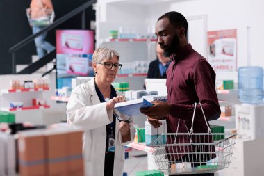 Afrika kökenli Amerikalı bir müşteri eczacıyla konuşuyor. Kardiyoloji hapları için yardım istiyor. Tıbbi broşürleri açıklayan bir işçi. Eczane çalışanı destek ve ilaç hizmeti sunuyor