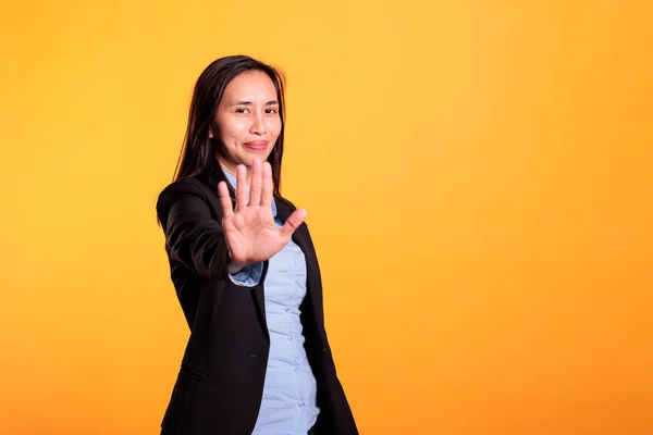 Negative filipino businesswoman doing stop gesture with hand in front of camera, posing in studio over yellow background. Serious woman showing refusal gesture, expressing rejection.