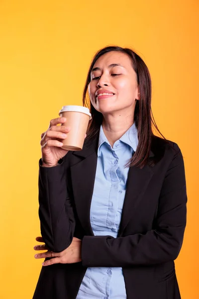 满意的亚洲模特儿拿着一杯咖啡喝着热饮料 享受着闲暇时间 然后开始在工作室拍照 年轻快乐的成年人 一头乌黑的长发 耸立在黄色的背景上 — 图库照片