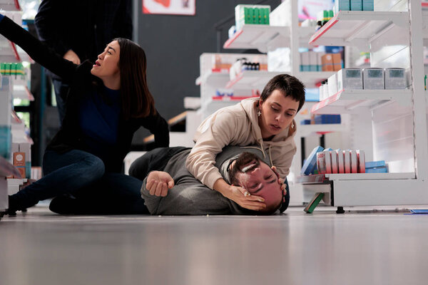 Аптека клиент держит человека голову во время приступа эпилепсии, оказание первой помощи. Разнообразные женщины, спасающие клиента аптеки во время эпилептического приступа в аптеке