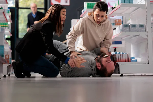Women standing on knees near unconscious man with epilepsy lying on pharmacy retail store floor. Young person suffering from foaming ar mouth and convulsions neurological disorder symptoms