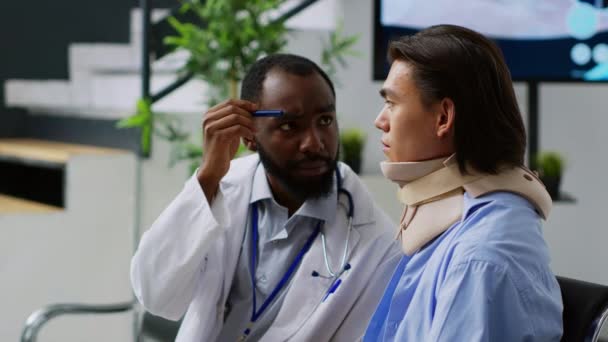 アフリカ系アメリカ人医師と脳損傷検査を行い 脳震盪をチェックし 医学的予約を持つ子宮頸椎のアジアの患者 病院のロビーで首の骨折や骨折をした人 — ストック動画