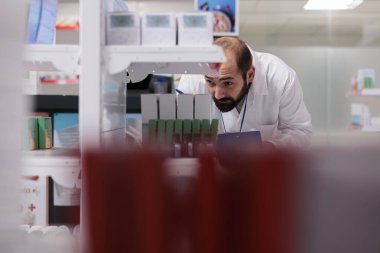 Eczacı ilaç paketleriyle dolu raflara bakıyor. Evraklara vitamin isimleri yazıyor, eczanede çalışıyor. Eczane çalışanı, doğruluğu sağlamak için verilen emirleri incelemekten sorumludur..