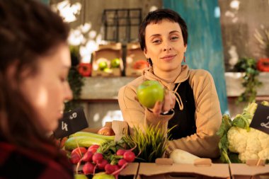 Marketin arkasında elinde yeşil elma tutan ve kameraya gülümseyen sağlıklı organik ürünlerle duran hoş, genç bir kadın çiftçi. Taze meyve ve sebze satan bir kadın.