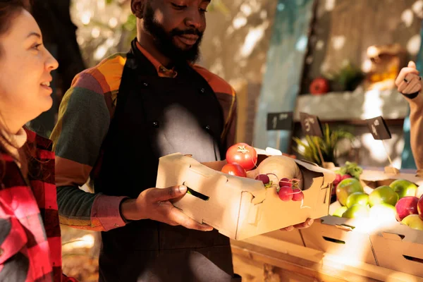 年轻的非洲裔美国男性农民在农贸市场上向顾客提供装满新鲜有机蔬菜的盒子 向消费者出售健康的当地种植的农场食品的小企业主 有选择的重点 — 图库照片