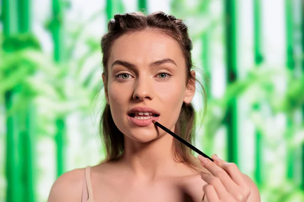 年轻模特儿用画笔画嘴唇 迷人的女人 拥有健康的新鲜肌肤 涂上唇膏 每天都在化妆 并在竹子背景下看着相机 — 图库照片