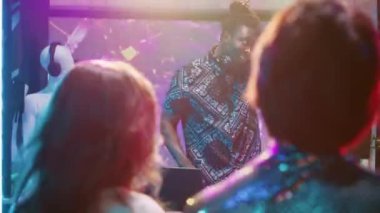 Erkek DJ partide ses karıştırma istasyonunu kullanıyor, modern müziği dans pistine koyuyor. Gençler sahne ışıkları ve canlı performansla eğleniyor. El kamerasıyla..