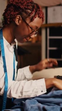 Dikey video terzisi, dikiş makinesi kullanarak iş yerindeki moda ürünlerini tasarlıyor. El işi aletleri ve aletlerle çalışan profesyonel bir terzi, özel dikim kıyafetler üretiyor..