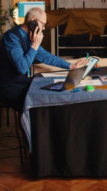 Dikey video eski tasarımcı atölyedeki müşteriyi arıyor, telefonda ölçümler ve kumaşlar hakkında konuşuyor. Özel dikim giyim markası üzerinde çalışan kıdemli bir terzi. El bilgisayarı