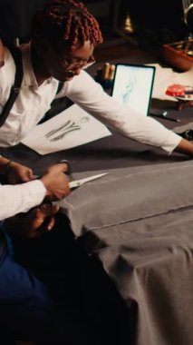 Dikey Afrikalı Amerikalı terziler elbise kesiyor, özel dikim kıyafetler yaratmak için takım halinde çalışıyorlar. Atölyedeki terzilik araçlarını kullanan zanaatkar ve asistan, dikiş işine hazırlanıyor