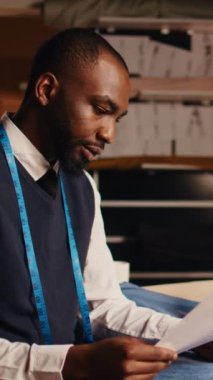 Dikey video Afrikalı Amerikalı terzi atölyedeki tasarımları karşılaştırmak için dizüstü bilgisayar ve kağıtları kullanarak çizimleri kontrol ediyor. Terzi, giyim eşyası modellerine bakıyor. El kamerasıyla.