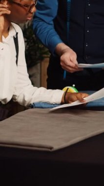 Dikey video terzisi ve çırak atölyedeki çizimleri inceliyor, stajyer giysi yaratmak için el sanatları öğreniyor. Tecrübeli tasarımcı terziye dikiş becerilerini anlatıyor.