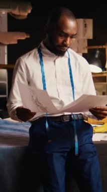 Dikey video Terzisi taslakları ve taslakları inceliyor, boynunda şerit var. Profesyonel moda tasarımcısı lüks giyim ürünleri ve zanaatkarlık yapmak için modeller arıyor..