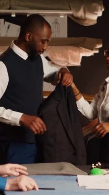 Atölyede takım elbise ceketi deneyen genç müşterinin dikey videosu. Doğru ölçümlere uyduğundan emin olmak için. Deneyimli terzi ve asistan, özel yapım spor ceket tasarlayıp atölyeye sığdırıyor..