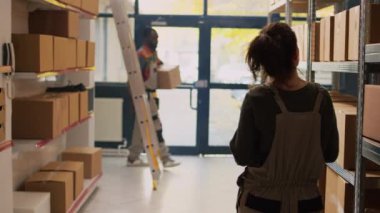 Depo malları, ambardaki ürünlerin listesini inceliyor. Raflarda paketlerle birlikte tedarik zinciri ve envanter üzerinde çalışıyor. Sipariş sevkiyatından önce malları inceleyen genç bir kadın..