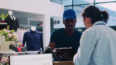 Asyalı çalışan dağıtım raporunu imzalıyor, alışveriş merkezindeki Afrikalı Amerikan kuryesiyle teslimat detaylarını tartışıyor. Modern butikte sevkiyat için çevrimiçi sipariş hazırlayan yönetici