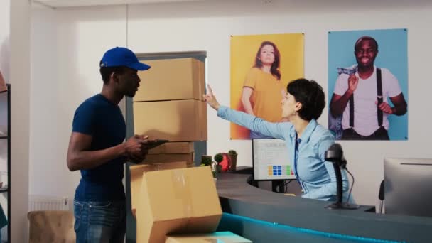 アフリカ系アメリカ人の配達員は 労働者にタブレットコンピュータの配布報告書に署名するよう求めており ショッピングモールの顧客出荷の詳細を議論している 現代的なブティックでのパッケージの準備 — ストック動画