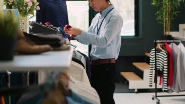 Asyalı işçi şık bir kravat takıyor, modern butikte aksesuarları rafa koyuyor. Perakende satış elemanı mağazayı açılışa hazırlıyor, alışveriş merkezinde şık kıyafetlerle çalışıyor. Moda konsepti