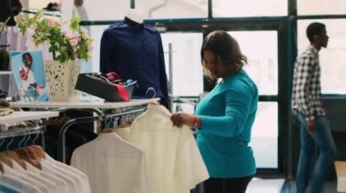 Beyaz gömlek için alışveriş yapan Afrikalı Amerikalı müşteriler, modern butikte kumaş parçalarını kontrol ediyorlar. Alışverişkolik kadın yeni moda koleksiyonu alıyor, giyim mağazasında günlük kıyafetlere bakıyor.