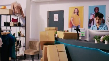 Afro-Amerikan kurye müdürle paket dağıtımını tartışıyor, nakliye raporunu modern butikte gösteriyor. Çevrimiçi emirlerle çalışan şık bir çalışan, teslimat için kutular hazırlıyor.