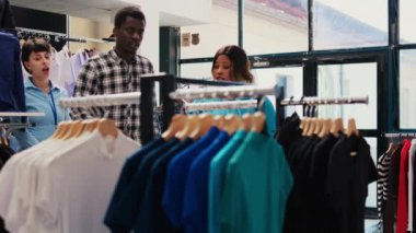 Çifte siyah gömlek gösteren şık bir çalışan, modern butikte yeni moda koleksiyonunu kontrol ediyor. Afro-Amerikalı müşteriler giyim mağazasından moda ürünler ve aksesuarlar alıyorlar.