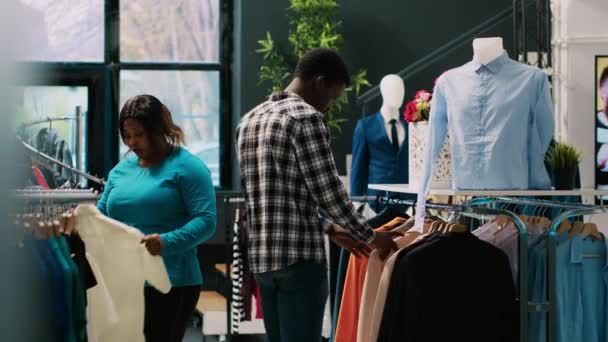 非洲裔美国人夫妇看着时髦的衣服 在现代精品店里买休闲装 愉快的顾客检查服装材料 为新衣橱购买时尚商品 — 图库视频影像