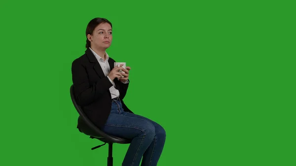 椅子にコーヒーカップを持っている白人実業家の側のビュー 緑の背景を持つスタジオに座って 女性マネージャーは カメラでカフェイン飲料やリフレッシュメントを飲む — ストック写真