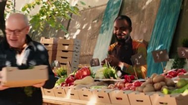 Team of farmers selling natural homegrown products to group of customers, talking to people about health benefits. Local vendors business owners giving box of fresh seasonal produce. Handheld shot.