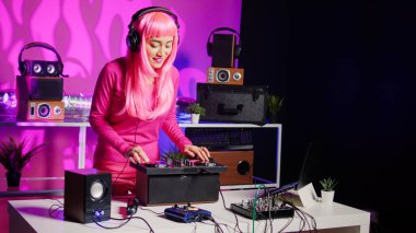 Smiling musician working as dj playing song at mixer console, mixing techno music with eletronic using audio equipment. Performer with pink hair having fun in performing in club at night time