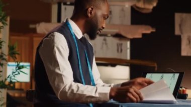 Tabletteki tasarımları inceleyen terzi atölyedeki kıyafetlerin çizimlerini karşılaştırıyor. Moda tasarımcısı terzilik aletleri ve aletleri kullanarak giyim markası üretiyor. El kamerasıyla..