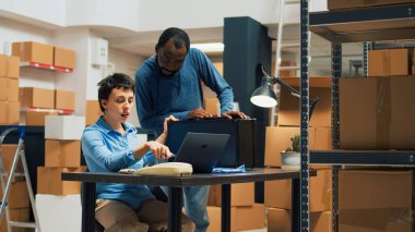 Diverse people working on merchandise shipping with laptop, preparing order in cardboard box. Man and woman putting products in packages, doing quality control before distribution. Tripod shot.