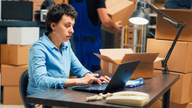 Business owner analyzing boxes with merchandise to ship order to clients, working on products distribution. Woman planning quality control and taking notes on laptop, supply chain management.